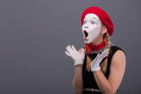 Waist-up portrait of young mime girl with red hat and red scarf looking aside with pleasant surprise, showing that she is happy to see something, isolated on grey background with copy placeの写真素材