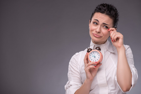 Close-up portrait of pretty girl with sad face crying and wiping her tears because she have no time, holding an alarm clock in her hand looking aside copy place isolated on grey backgroundの写真素材