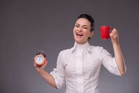 Waist- up portrait of happy girl holding an alarm clock and a red cup in her hand looking at the camera with pretty smile isolated on grey background with copy place concept of time managementの写真素材