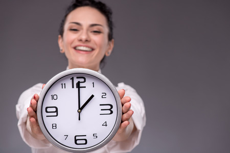 Waist-up portrait of beautiful smiling girl holding a big clock in her hands and looking at the camera, with selective focus on a clock isolated on grey background with copy placeの写真素材