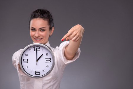 Waist-up portrait of beautiful smiling girl holding a big clock in her hands and looking at the camera, with selective focus on a clock isolated on grey background with copy placeの写真素材