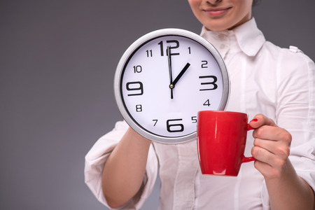 Close-up portrait with selective focus of a big clock and a red cup in hands of a young woman...