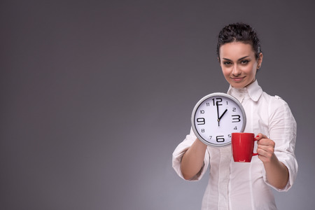 Waist-up portrait of a young beautiful girl holding a big clock and a red cup in her hands and happy...