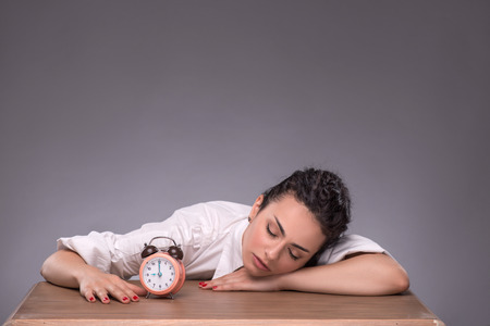 Waist-up portrait of young girl sitting at the table with small alarm clock and holding her hands on...