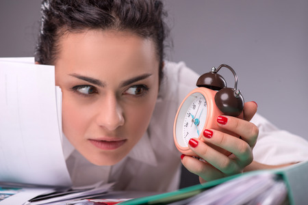 Close-portrait of confused girl sitting at the table with a pile of documents and looking with a fright at a clock, isolated on grey background with copy place, concept of time management at workの写真素材