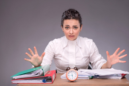 Waist-up portrait of girl sitting at the table with documents and an alarm clock, breeding her hands because she has no time, confused looking at you, isolated on grey background with copy placeの写真素材