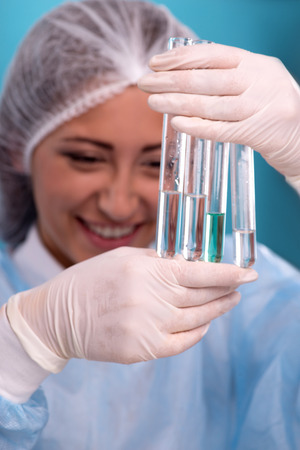 Close-up portrait of female scientist in overall holding in her hands four tubes with some substances in laboratory while doing some experiments isolated on blue background, with selective focus on a tubesの写真素材