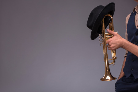 Close-up picture of a trumpet with a black hat on it in hands of a jazz man in a suit isolated on grey background with copy place, concept of jazz musicの写真素材