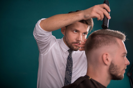 hairdresser  cuts   hair  with hair clipper  on crown of handsome satisfied  client  in  professional  hairdressing salonの写真素材
