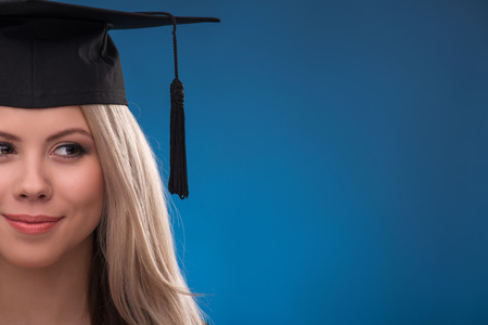 Portrait of beautiful happy blond girl in black cloak and master hat on blue background smiling looking aside  close up with copy placeの写真素材