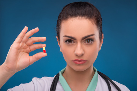 Portrait of beautiful woman doctor on blue background with stethoscope looking at camera holding with fingers red white tabletの写真素材