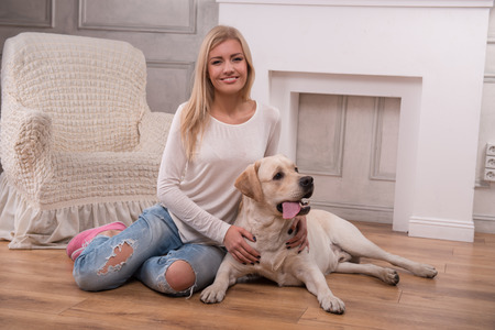 Beautiful  slender blond girl in jeans and beige knitted jacket  sitting on floor  with Labrador Retriever smiling looking at camera full lengthの写真素材