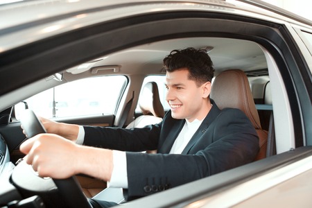 Confident drive. Portrait of a smiling handsome man in suit sitting on drivers place in car and...