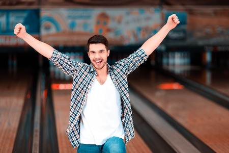 Bowling master. Cheerful young man raising his hands high above the head and smiling at camera while standing against bowling alleys after the strikeの写真素材