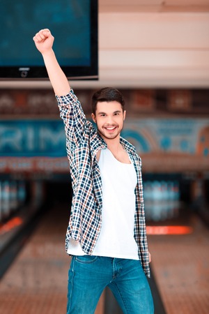 Bowling master. Cheerful young man raising his hands high above the head and smiling at camera while standing against bowling alleys after the strikeの写真素材