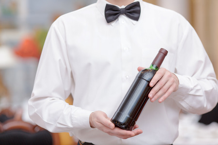 Confident waiter. Cropped image of handsome young waiter in shirt and bow tie holding a wine bottle in the luxury restaurantの写真素材