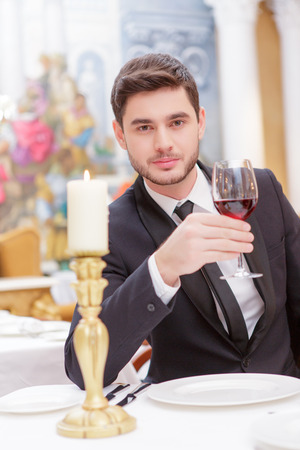 Making a toast. Handsome young man in suit raising his glass of wine while making a toast in luxury restaurantの写真素材