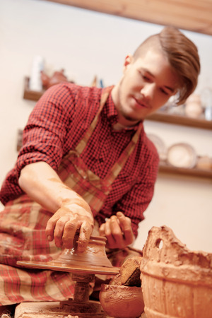 Clay modelling. Close-up of a young man in casual wear and apron shaping a clay pot in studioの写真素材
