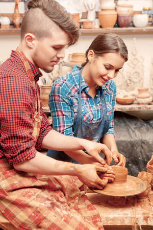 Excitement of art. Young pretty couple in aprons creating a ceramic item with a pottery wheelの写真素材