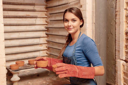 Interesting hobby. Young smiling girl wearing gloves puts clay pots to the shelf so that they could get dryの写真素材