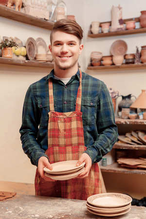 Best self-made crockery. Young handsome guy holding ceramic plates made in clay studioの写真素材