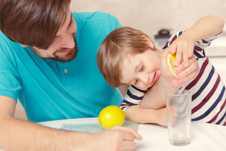 Home lemonade. Young father and his son squeezing lemon to the glass of water at home kitchenの写真素材