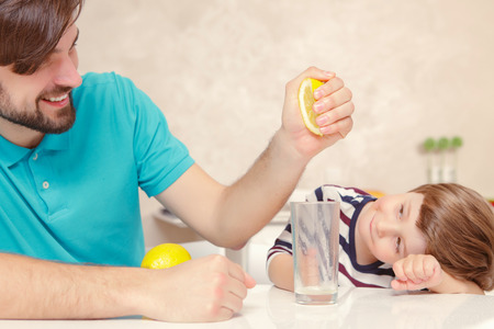 Home lemonade. Young father and his son squeezing lemon to the glass of water at home kitchenの写真素材