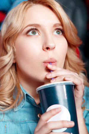 Rapturous girl. Young blond woman enthusiastically watching film and drinking coke in cinemaの写真素材