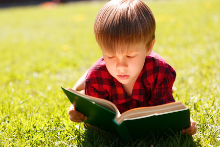 So catching. Close up of young boy lying on green grass and reading big book.の写真素材