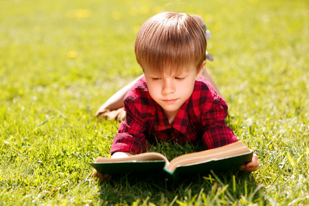 I love reading. Portrait of cute little boy lying on green grass and reading book.の写真素材