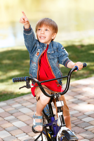 Little cute smiling boy riding bicycle in park and pointing upwards with his index finger.の写真素材