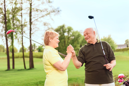 One team. Smiling senior man and woman standing on course holding golf clubs and their hands togetherの写真素材