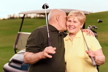 Showing love. Senior man with golf club giving kiss on cheek to his wife on background of cart on course.の写真素材
