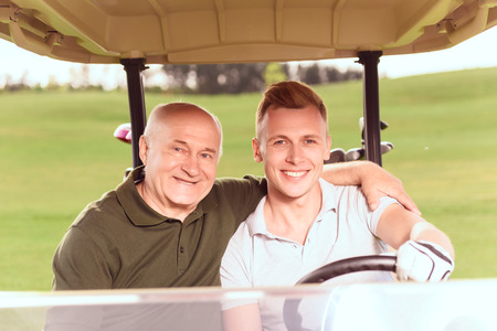 Supporting embrace. Smiling young and senior men driving in cart on course, old man embracing young one.の写真素材