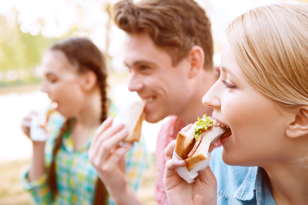 Biting off. Group of young people sitting on cover and eating sandwiches during picnicの写真素材