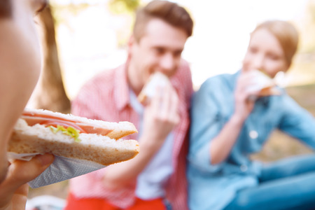 First bite.  Close up of young girl eating sandwich on background of another people during picnicの写真素材
