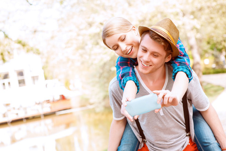 Out of way. Handsome man with hat carrying his girlfriend on his back that is doing selfie on background of lake.の写真素材