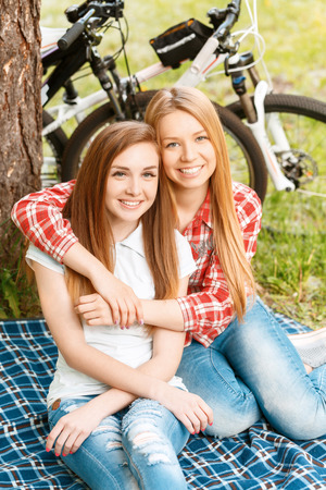 Two young beautiful girls with long hair sitting on a blue checkered mat under a tree smiling happily and hugging, their bikes standing near in a green park, top viewの写真素材
