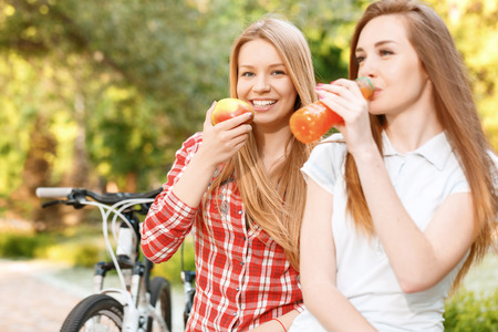 Beautiful young girl with long brown hair sitting in a park and drinking fruit juice with her blond friend smiling and holding a big tasty apple their sport bikes standing near, selective focusの写真素材