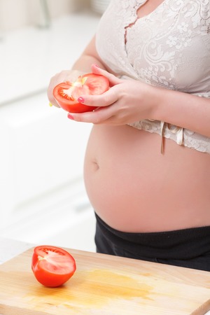 Bright cooking. Pregnant young woman cleaving tomato while cooking healthy salad in the kitchen.の写真素材