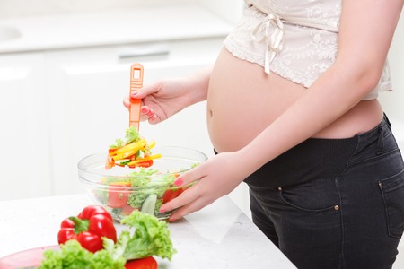 Tasty cooking. Pregnant young  woman mixing colorful vegetable salad while preparing  nutritional food in the kitchenの写真素材
