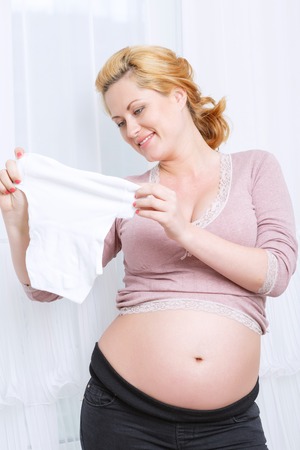 Expecting kid. Young pregnant woman stretching baby clothes while standing near the window.の写真素材
