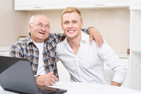 Friendly embrace. Grandson and his grandpa sitting together in kitchen with laptop and embracing.