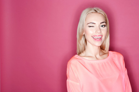 Giving wink. Portrait of astonished young blond-haired woman standing on isolated pink background and winking.の写真素材