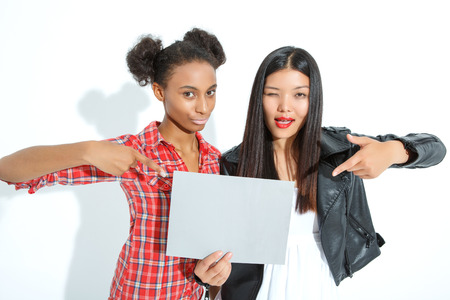 Look here. Nice agreeable young ladies holding sheet of paper and pointing it with index fingers while standing isolated on white background.の写真素材