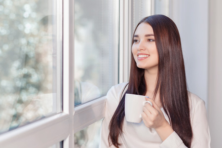 Time to relax. Vivacious  nice business woman holding cup and standing near window while reveling in...