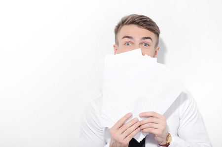 Hiding from you. Young-looking  attractive businessman standing on white isolated background holding papers and covering his face with them.の写真素材