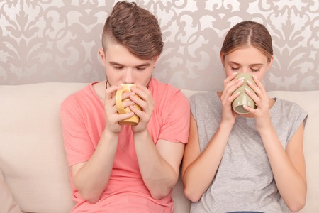 Revel in great aroma. Contented pleasant young couple sitting on the couch and holding cups while rejoicing gin drinking tea.の写真素材