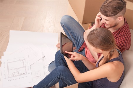 Prepare for living. Pleasant young wife sitting near settee and holding laptop while pointing it to...