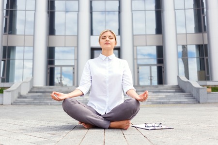 In harmony with mid. Pleasant beautiful young businesswoman sitting on the ground near office building and practicing yoga while feeling delightedの写真素材